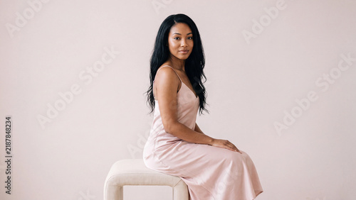 Black woman with long dark hair wearing a light pink slip dress sitting gracefully on a cream colored stool in a bright minimal studio space.