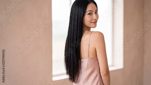Model with long dark hair posing gracefully in front of a window inside a minimalist room with warm neutral walls and soft daylight illuminating her face.