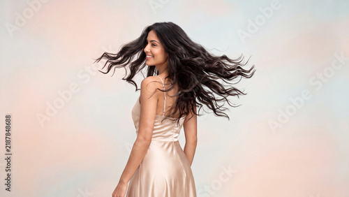 Brunette woman with long flowing hair tossing her head back dynamically while wearing an elegant satin evening gown against a soft pastel studio backdrop.