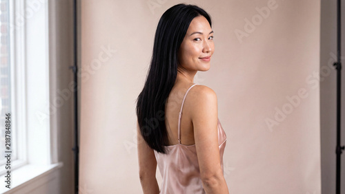 Asian woman with long straight black hair smiling over her shoulder while wearing a pink silk slip dress inside a bright studio with a neutral background.