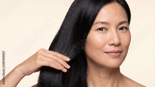 Mature woman gently touches her long glossy black hair with a serene expression while displaying healthy and well-maintained strands against a neutral plain background.