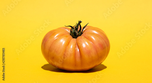 A single fresh heirloom tomato rests centered on a vibrant yellow background featuring bright and crisp studio lighting.