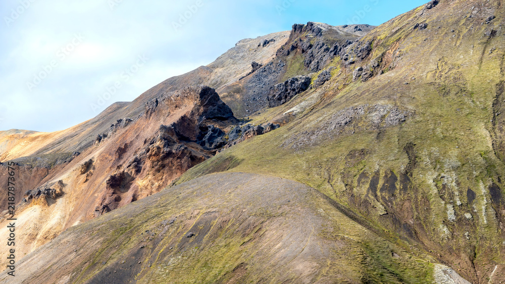 Obraz premium Colorful layered mountains with moss-covered slopes and dark rock formations create a dramatic landscape in Landmannalaugar, Icelandic highlands.