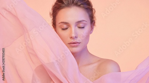 A woman with closed eyes resting her head against flowing pink fabric in a soft pastel studio setting with closed rests
