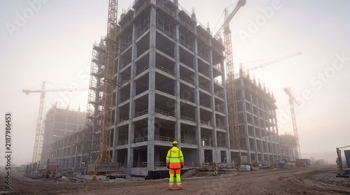 Construction worker wearing bright safety gear standing before unfinished concrete building frames during early morning fog