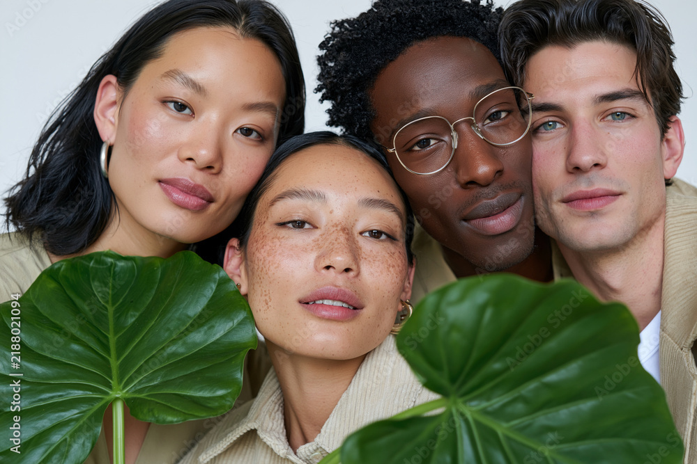 Fototapeta premium Four diverse individuals pose closely together. Large, vibrant green leaves frame their faces. They look directly at the camera with calm, natural expressions. Soft lighting enhances their skin tones
