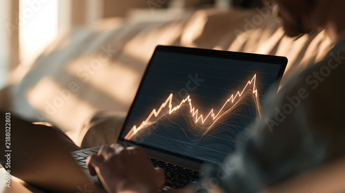 Person using a laptop to monitor glowing financial market data charts while sitting at home with screen illuminating dark indoor environment