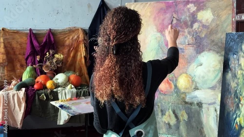 Teenage female artist in college painting class, art studio, art workshop. She paints still life on canvas with oil paints, holds brush, palette.