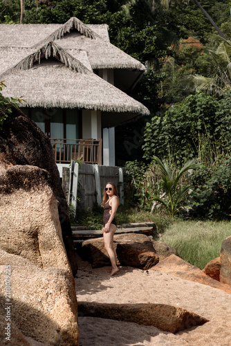 Bild auf Leinwand Woman in a swimsuit enjoying a tropical beach beside large rocks and a traditional bungalow, representing Thailand travel, resort vacation and tropical lifestyle