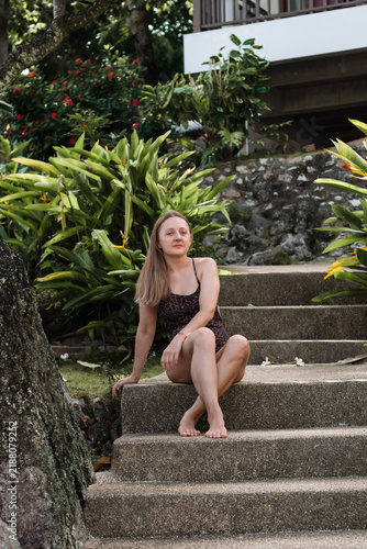 Obraz na plátně Woman in a swimsuit relaxing on stone steps surrounded by lush tropical greenery, offering a natural travel and resort vacation lifestyle concept