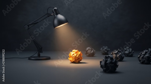 A desk lamp shines a bright light on a single orange crumpled paper ball amidst many dark ones on a dark surface.