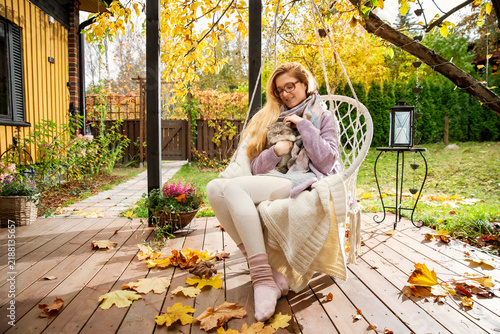 A woman in her late 30s relaxes in a hanging chair on an autumn patio, holding her cat in a cozy hygge moment.