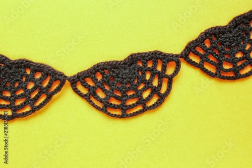 A top-down view of a black crocheted lace border with a semi-circular openwork pattern resembling a spider web, arranged horizontally across a bright solid yellow paper background with copy space