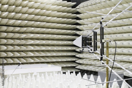 An indoor view of an anechoic chamber for electromagnetic compatibility testing, featuring walls lined with acoustic foam pyramids, a log-periodic antenna, and a white testing table layout