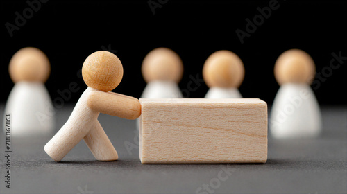 Wooden figure pushing block with blurred figures in background, teamwork concept