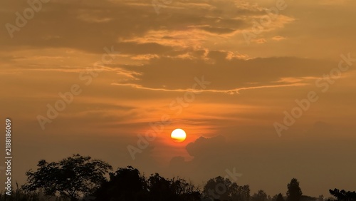 Golden sun setting behind clouds over a dark treeline horizon