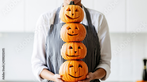 a cook holding a vertical stack of three handmade jack-o-lanterns in front of a striped apron, carved triangular eyes and smiling mouths forming a whimsical pumpkin tower in a brig