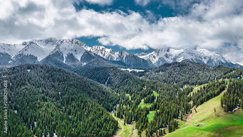 Fotografía Aerial view of snow-capped mountain ranges under bright sunlight, with dense green pine forest and white clouds over Xinjiang, China