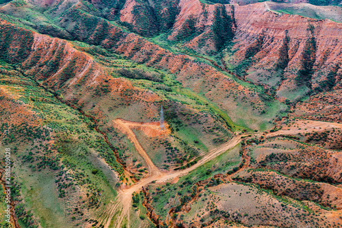 фотографія High angle view of rugged red and green landforms in Xinjiang, displaying layered rock erosion, dirt tracks, and scattered vegetation under soft daylight