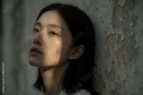 Close-up portrait of a young Asian woman leaning against a peeling wall in natural light