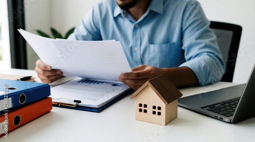 Man reviews property documents at desk with laptop. Tiny house model sits near papers and binders. He analyzes contract for home investment or mortgage loan.