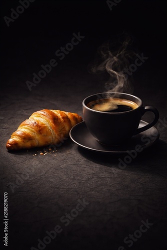 Steaming hot espresso in a black cup with a golden croissant on a dark table.