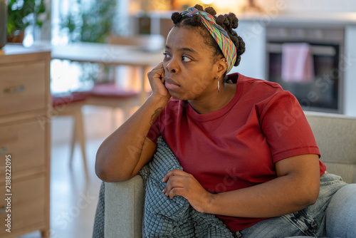 Принти на полотні Tired african american woman rests head on hand while sitting alone at home, looking tired, bored, unmotivated