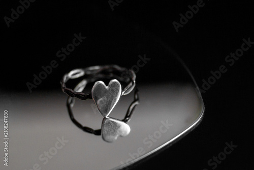 Konstfotografi A silver ring with two heart shapes intertwined, resting on a reflective surface