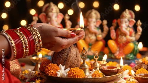 Woman holding burning clay diya with traditional red bangles over brass puja thali containing coconut, marigold flowers, and bell for Diwali ... See More