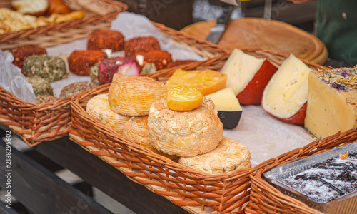 Assortment of artisan craft cheeses displayed in wicker baskets at an outdoor farmers market counter. Assorted goat and cow milk cheeses with herbs, spices, and crusts