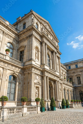 A magnificent exterior view of a grand historic palace museum facade featuring ornate classical stone architecture, tall columns, intricate statues and a cobblestone courtyard under a clear blue sunny