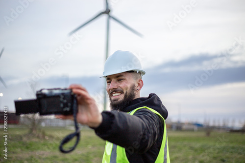 Happy engineer in a hard hat and safety vest is smiling while making a video at a wind turbine site