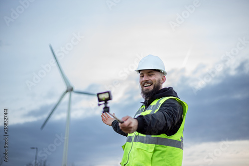 Engineer recording vlog on renewable energy with action camera and selfie stick near wind turbine