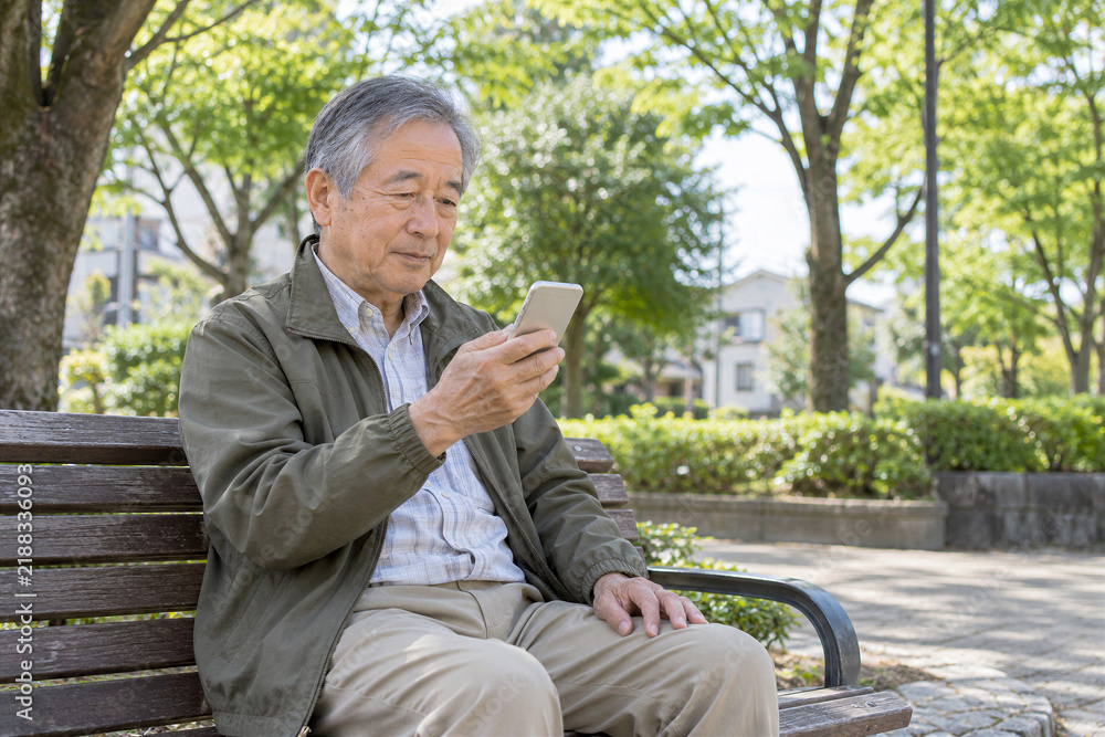 Obraz premium 近所の公園のベンチでスマートフォンを使う日本人シニア男性