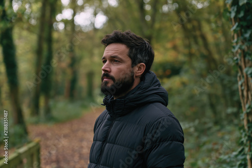 A thoughtful man wears a puffer jacket while reflecting amidst a forest backdrop, perfectly capturing a blend of nature, introspection, and personal style in a tranquil setting.