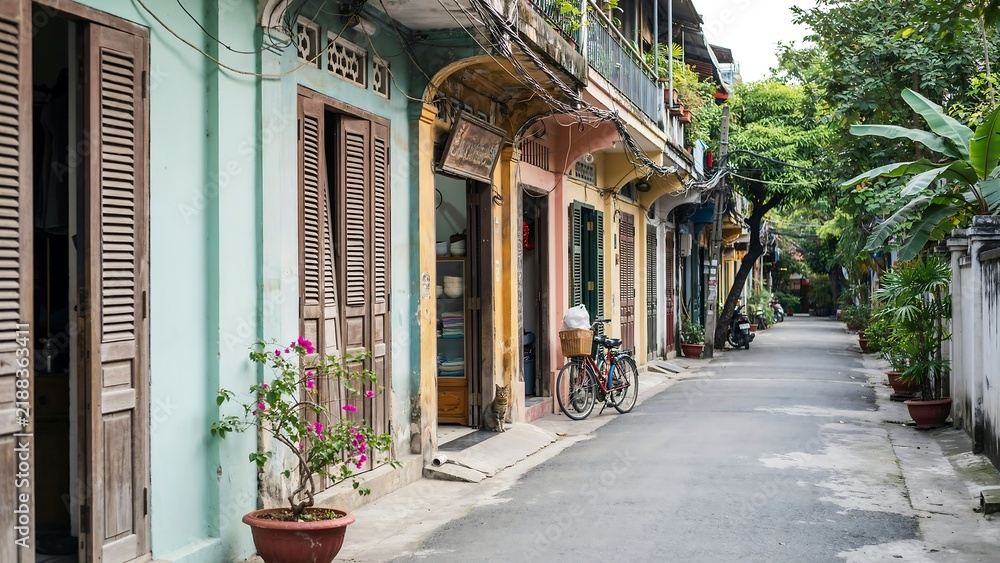 Fototapeta premium Narrow street in an old town with colorful colonial buildings, wooden shutters, potted flowers, and a bicycle parked against a textured wall