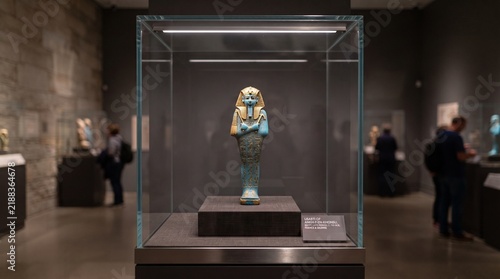 Ancient Egyptian Usabti artifact displayed in a museum exhibit behind glass with visitors in the background