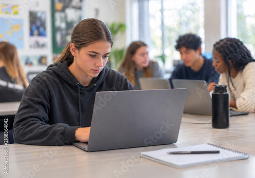 Focused female high school student working on a laptop in a modern classroom with blurred diverse students in the  background.