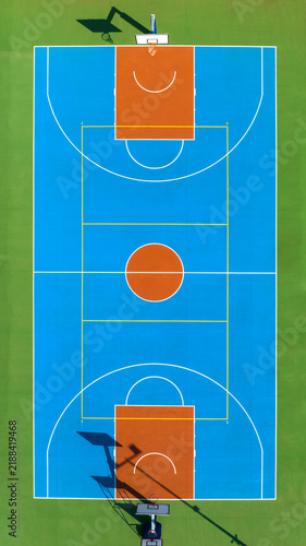 Vertical aerial view of a vibrant blue and orange outdoor sports court set on green grass, featuring white field markings and basketball hoops casting long shadows in the sunlight. Zenithal shot.