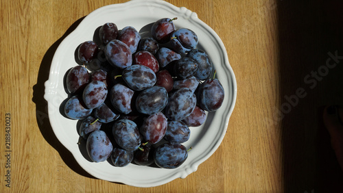 Photography Freshly picked purple plums with a dusty blue-grey bloom on the skin, piled in a white ceramic bowl on a rustic wooden table, homegrown harvest for recipe features and seasonal eating content