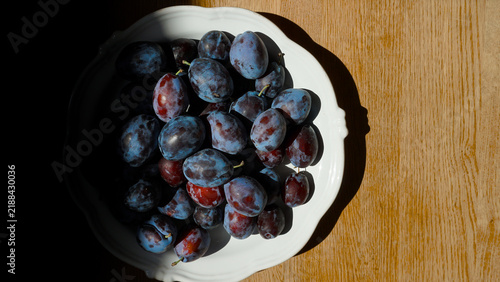 фотографія Freshly picked purple plums with a dusty blue-grey bloom on the skin, piled in a white ceramic bowl on a rustic wooden table, homegrown harvest for recipe features and seasonal eating content