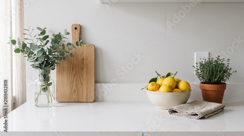 Modern minimalist kitchen counter with fresh lemons in ceramic bowl, wooden cutting board, eucalyptus branches in glass vase, potted thyme herb, bright clean interior design