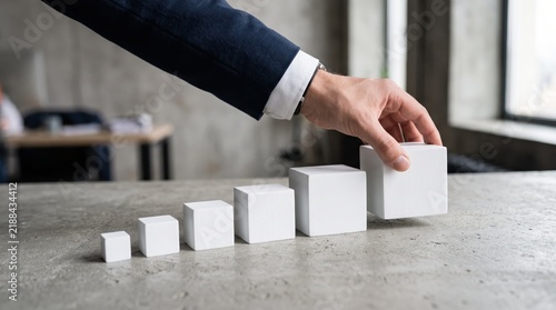 Businessman arranging white cubes in ascending order on concrete table, business growth concept, step by step strategy, progress, development, scaling up, success ladder, building