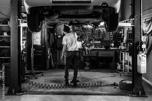 Black and white photo of a mechanic inspecting the underside of a raised car in a busy auto repair garage with tools, equipment, and workshop details visible