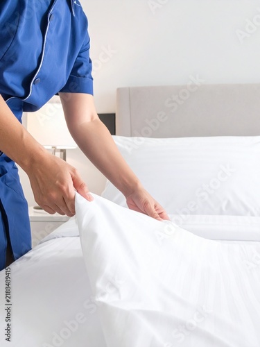 A person in a blue uniform making a bed with fresh white linen in a hotel room.