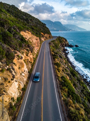An aerial view of a car driving on a scenic coastal road winding along a cliffside with the ocean on one side and green hills on the other under a cloudy sky.