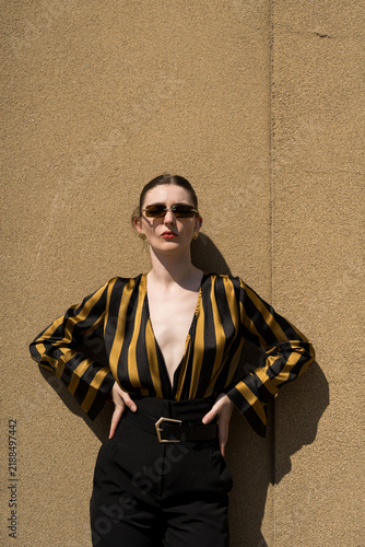Portrait of attractive young caucasian woman wearing yellow and black striped blouse and sunglasses, standing near a wall on a warm, sunny summer day. Looking at camera. Fashion, style, beauty.