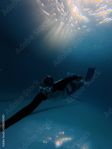 Freediver glides horizontally underwater in deep pool beneath bright sunbeams