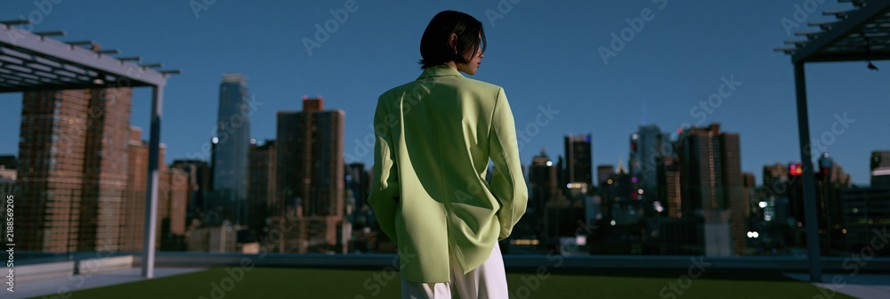 Fototapeta premium Rear view of a person wearing a chartreuse jacket on a rooftop terrace with a city skyline at twilight for urban fashion wallpaper and banner