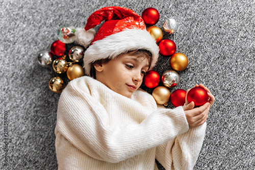 Cute boy in a Santa hat holding a Christmas ball on a grey carpet. Festive holiday season childhood concept.